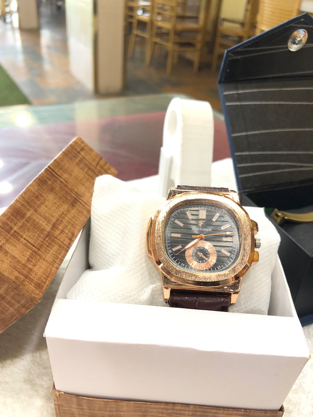 Luxury watch with rose gold case and black dial in a white box on a glass table.
