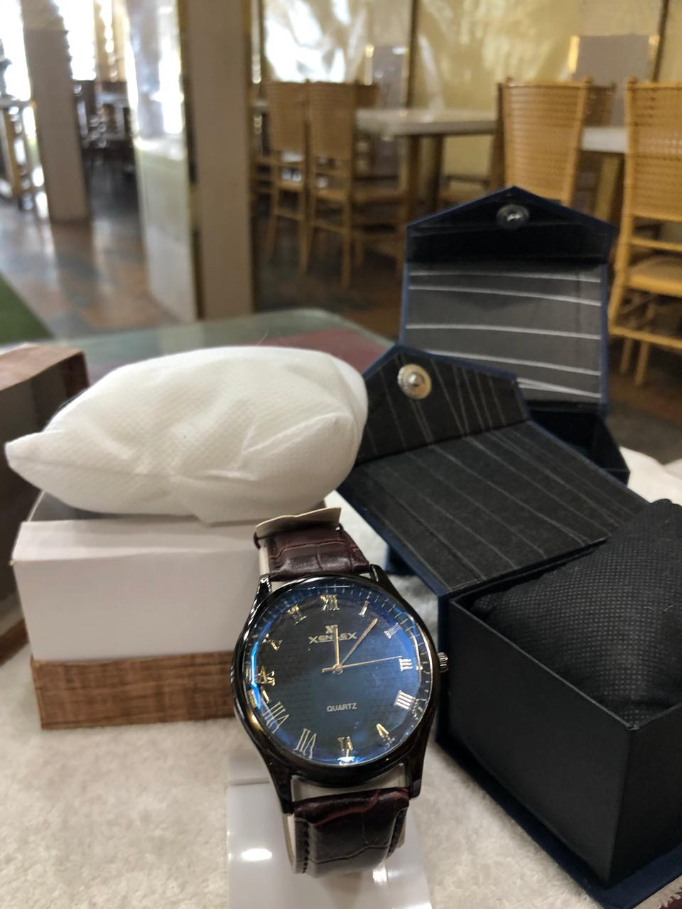 Wristwatch with blue face and brown leather strap on a reflective surface, with a blurred indoor background.