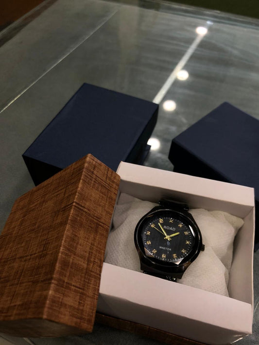 Black watch with gold accents in a white box, surrounded by wooden and dark blue boxes on a reflective surface.